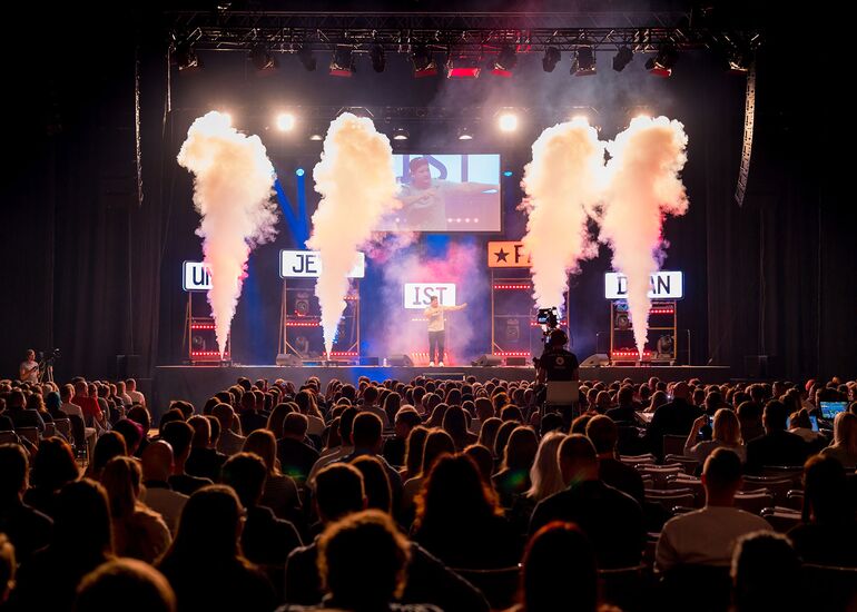 Aus dem Publikum ist Comedian Chris Tall auf der Bühne der GETEC-Arena zu sehen. Neben ihm sieht man Bühnenrauchfontänen und im Hintergrund Leuchtschilder mit Wörtern .