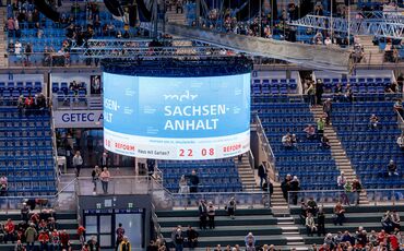 LED Zylinder mit Werbung für MDR Sachsen-Anhalt hängt an der Decke der GETEC-Arena Magdeburg. Im Hintergrund ist die blaue Reihenbestuhlung der Ränge zu sehen.